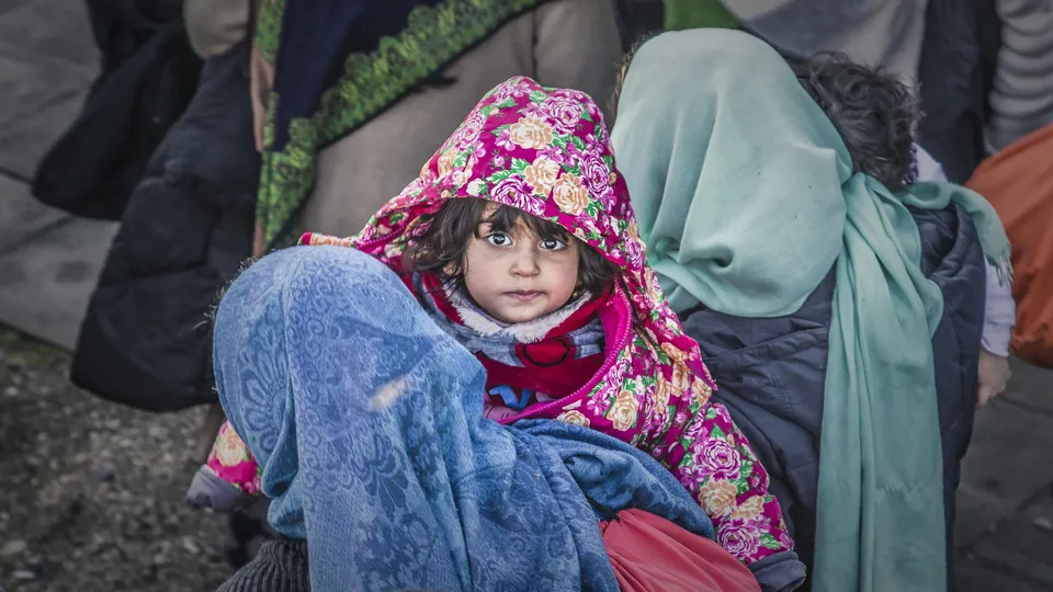 A small girl in a pink floral hood looks up from her mother's arms in a crowd of refugees
